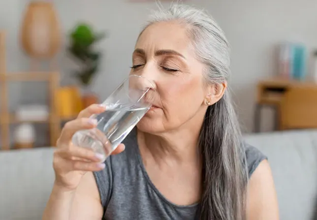 A woman drinks water from a glass. She is sitting on a sofa in a living room.