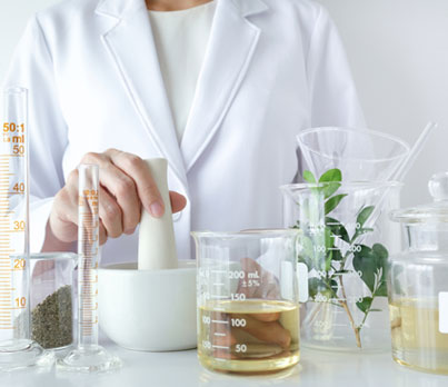 A mortar and pestle are being used by a scientist wearing a white lab coat. Various beakers, graduated cylinders, and flasks containing liquids and plant matter are present on a white surface.