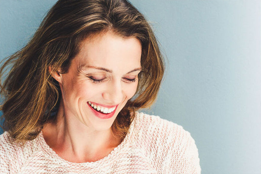 A woman with shoulder-length brown hair, wearing a red sweater, smiles gently while standing against a rough, light-grey stone wall.