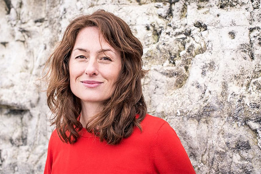 A woman with shoulder-length brown hair, wearing a red sweater, smiles gently while standing against a rough, light-grey stone wall.