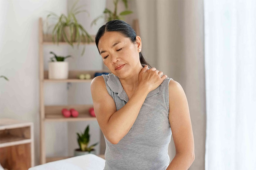 A woman massages her shoulder, experiencing pain; she is in a bright, minimalist room.