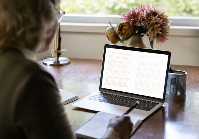 A person writes in a notebook while referencing a laptop displaying text; the scene is set at a desk near a window.