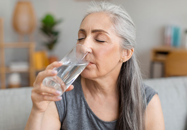 A woman drinks water from a glass. She is sitting on a sofa in a living room.