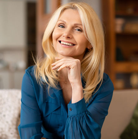 A blond woman, smiling, rests her chin on her hand. She wears a teal blouse. The background suggests a home living room.