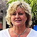 A woman with short, curly blond hair is looking at the camera, wearing a white shirt and a necklace, outdoors amidst green foliage.