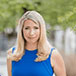 A woman with shoulder-length blonde hair is smiling subtly, wearing a sleeveless royal blue top. She is posed outdoors against a softly blurred background of green foliage.